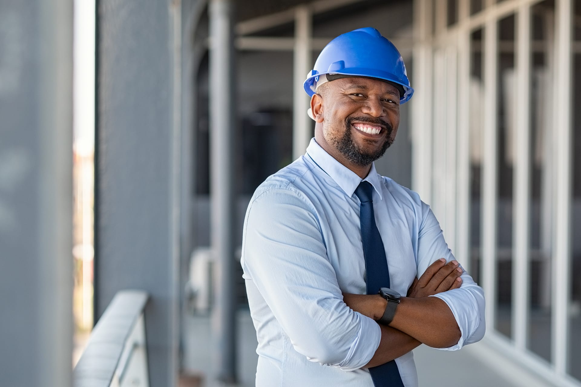 Portrait of african american man architect at building site with folded arms looking at camera. Confident construction manager in formal clothing wearing blue hardhat. Successful mature civil engineer at construction site with copy space.